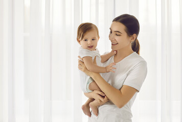 Portrait in bright room of young loving mother holding her beautiful six month old daughter. Mom...