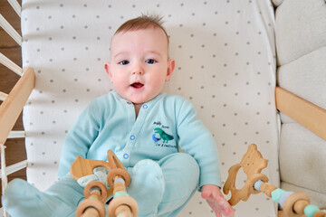 Happy infant baby is playing with wooden hanging toys lying in bed. Smiling funny child in...