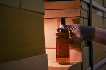 A man gloved hand holds a bottle of alcohol in a warehouse with boxes for delivery to customers and...