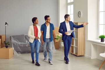 Fototapeta premium Realtor or real estate agent giving house tour to future buyers. Young black couple planning to buy new home. Boyfriend and girlfriend or husband and wife looking at modern, big, spacious apartment