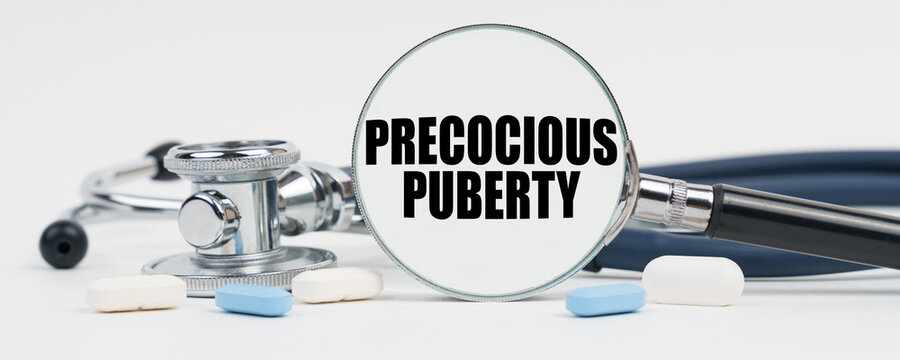 On a white surface are pills, a stethoscope and a magnifying glass inside which is written - Precocious puberty