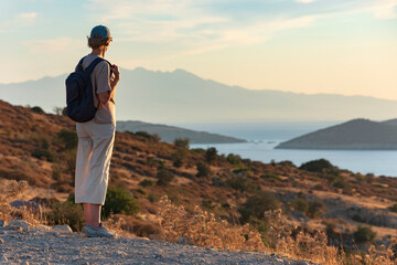 A middle-aged woman, a traveler with a backpack, standing on top of a mountain at sunset next to the sea. A tourist enjoys the panorama of the beautiful bay landscape. Turkey, Bodrum.