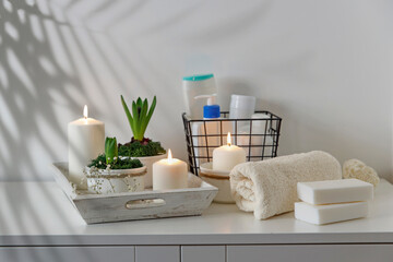 Bathroom decor. Unblown hyacinths with burning candles on a wooden vintage tray. Palm tree shadow on the wall. Shampoos and cream in a metal basket. Rolled white towel and two bars of soap