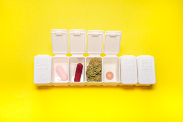 White pillbox with a big cannabis bud and different colorful pills on yellow background. Concept of medical marijuana and CBD, natural medicines..