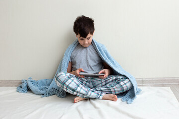 A brunette boy, a teenager in gray pajamas, on the floor, smiles, uses a digital tablet