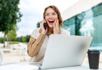 Office employee talking on cell phone with smile sitting on terrace in cafe with coffee and laptop