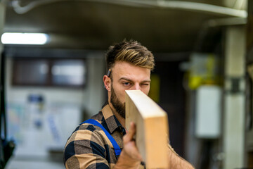 Young handsome caucasian carpenter checking the wooden board in old way - obrazy, fototapety, plakaty
