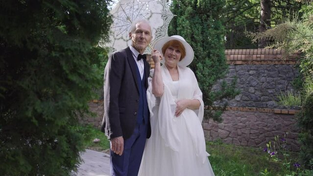 Romantic Loving Senior Couple In Wedding Dress And Suit Posing With Sun Umbrella In Slow Motion Outdoors. Portrait Of Happy Caucasian Man And Woman Getting Married On Summer Spring Day Smiling