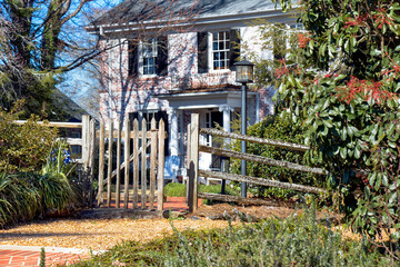 A brick and pea-gravel path, a weathered split rail fence with a wooden picket gate, and tall evergreen shrubs make an inviting entrance to a stately brick colonial house. Focus on foreground.