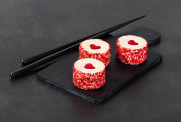 Festive creamy white chocolate mousse cakes with a jelly heart, in the form of round sweet sushi, sprinkled with small pieces of freeze-dried strawberries on a serving board on a dark gray background