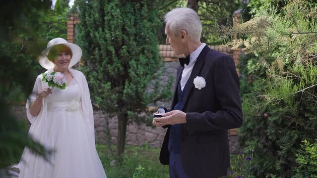 Happy proud senior man in wedding suit admiring rings and blurred woman in wedding dress at background turning looking at camera. Portrait of loving Caucasian groom posing outdoors in slow motion