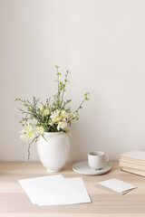 Fototapeta na wymiar Floral still life. Easter bouquet. Wite, yellow tulips, daffodils flowers. Green birch tree, blueberry branches in ceramic vase pot on wooden table. Vertical. Blank paper cards mockups, cup of coffee