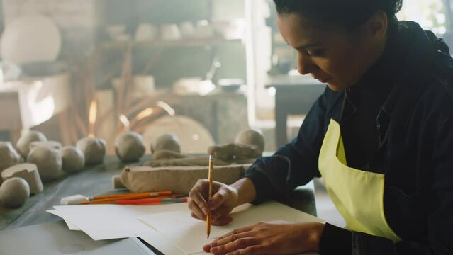 Young woman potter is sitting at table and working on new items collection from clay, making sketches of pots and vases on paper, close up, Slow motion.