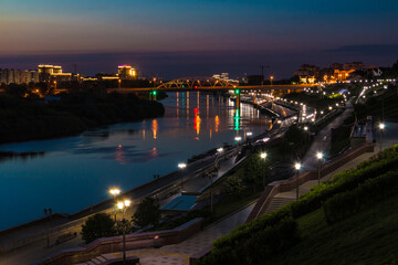 Fototapeta premium Beautiful view of the illuminated Embankment of Tura River and the Chelyuskintsev Bridge at dusk, Tyumen, Russia 