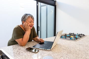 Senior woman 68 years old working on laptop and watching video in kitchen