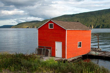 Fototapeta na wymiar An orange wooden boat shed with small glass windows and a single hung white door on the ocean. There are fish boxes stacked near the entrance. The ocean and mountains are in the background. 
