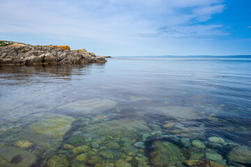 A wide cove or bay with hills covered in green trees under thick cloudy skies at a point. The sandy curved shaped beach has lush green grass and a row of red seaweed near the ocean's edge. 