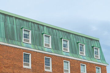 The corner view of a vintage brick building with a copper hip roof that has oxidized to a pale green outer layer, patina.  The bricks are small in a horizontal pattern. There are multiple windows. 