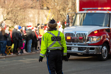 Police officer, wearing fluorescent clothing, standing in a crowded street. The officer is wearing a bulletproof vest with the word police on her back. A group of young people is in the background.