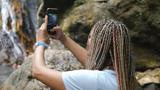 Female taking picture for blog standing in public park on nature waterfall background. Wonam with long braids hairstyle