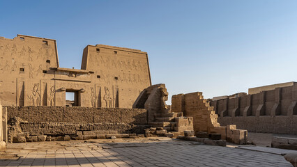 The Temple of Horus in Edfu against the blue sky. Huge carved images of the gods are visible on the high wall. Ruins are in the foreground. Egypt