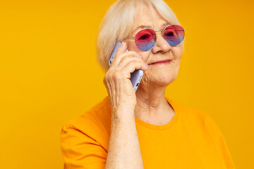 smiling elderly woman talking on the phone in yellow glasses yellow background