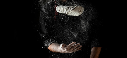 Naklejka na ściany i meble flying pizza dough with flour scattering in a freeze motion of a cloud of flour midair on black. Cook hands kneading dough. copy space