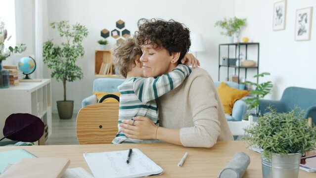 Little boy doing homework with mum hugging talking doing high-five indoors at home. Family relationship and domestic education concept.