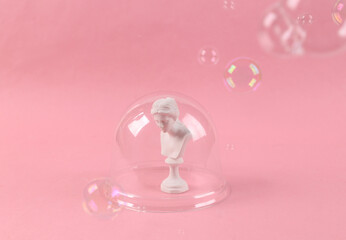 Creative art, minimal layout with a bust of venus under a transparent dome and soap bubbles on a pink background