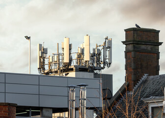 5g mobile phone radio mast transmitter at sunset in residential town centre with old 4g and 3g antennas also attached.  Buildings below with bright sky and city rooftops.