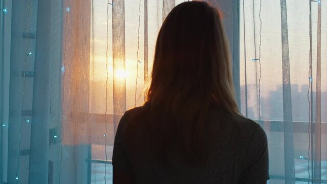 Young woman watches sunset trough the window
