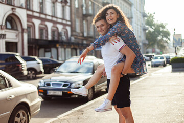 Fototapeta na wymiar Couple in love. Man carrying girl on his back in the street. Smiling man with beautiful young woman, ride piggyback, having fun together. Relationship concept.