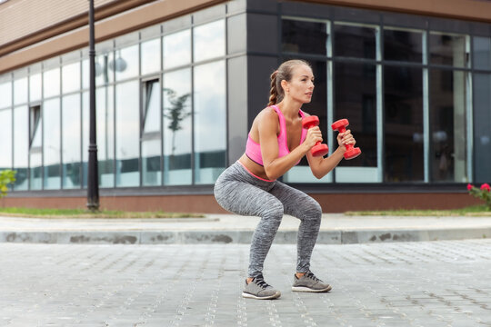 Active lifestyle concept. Fit woman in sportswear trains leg muscles with dumbbells in hands outdoors