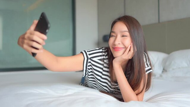 Portrait of young smiling asian woman in bed taking selfie photo and video video with smartphone front camera