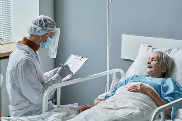 Young doctor in protective wear writing prescription in medical card while visiting senior woman at hospital ward