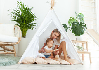 Mom and daughter are playing with a developing pyramid while sitting in wigwam in modern minimalistic light room with house plants © splitov27