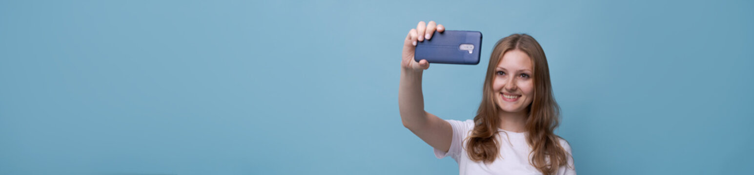 Young female vlogger with surprised face takes picture herself on phone while broadcasting on a blue background in a white t-shirt. Caucasian beautiful girl. Selfie concept story and blog