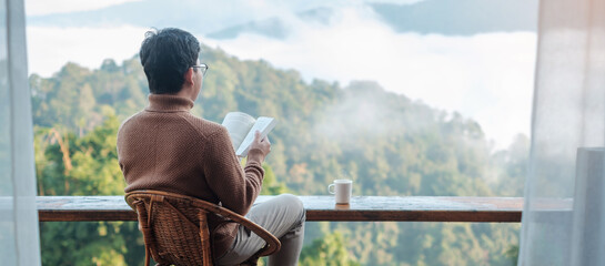 young man reading book near window and looking mountain view at countryside homestay in the morning sunrise. SoloTravel, journey, trip and relaxing concept