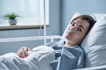 Portrait of young woman lying on bed with oxygen tube looking at camera while treating at hospital