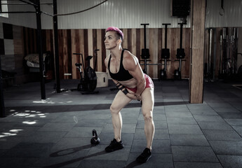 Fototapeta na wymiar Muscular extraordinary female athlete with short pink hair trains swing with heavy kettlebells. Functional, cross training in modern gym.