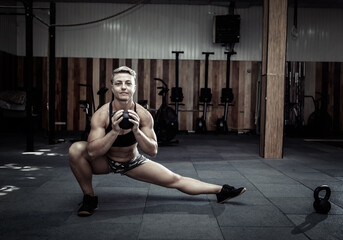Fototapeta na wymiar Muscular extraordinary female athlete with short pink hair trains side lunges with heavy kettlebells. Functional, cross training in modern gym.