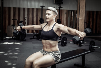 Fototapeta na wymiar Young athletic strong woman trains the muscles of the shoulder with dumbbells in her hands while sitting on a bench in a cross gym. Bodybuilding and Fitness