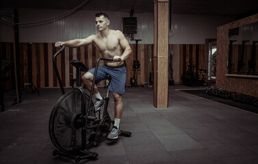 Fototapeta na wymiar Powerful muscular male athlete with a naked torso trains with an air bike in a modern gym. Functional training. Cardio exercise. Healthy lifestyle concept