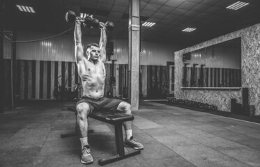 Powerful athletic man training shoulder muscles with heavy dumbbells doing overhead while sitting on a bench in gym. Healthy lifestyle concept
