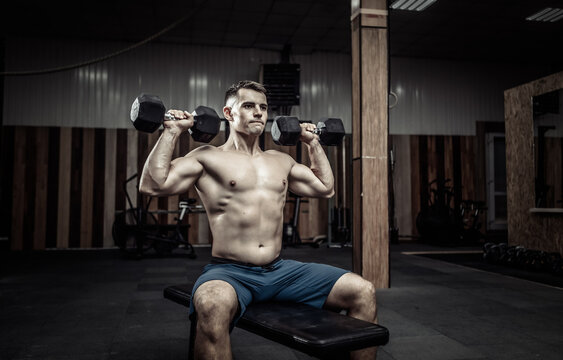 Powerful athletic man training shoulder muscles with heavy dumbbells while sitting on bench in gym. Healthy lifestyle concept