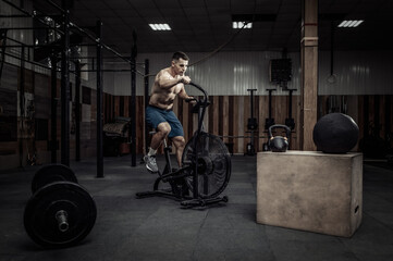 Fototapeta na wymiar Powerful muscular male athlete with a naked torso trains with an air bike in a modern health club. Functional, cross fit training. Cardio exercise. Healthy lifestyle concept