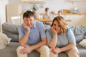 Tired parents suffer from headaches, sit on couch, while their daughter and two sons run in the kitchen and scream, dad and mom feel exhausted by noisy children. Disobedient children concept