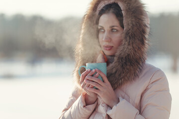 Winter image of a young woman in a down jacket with fur, looking into the distance. In the hands of a blue cup with hot tea. The hot steam coming from the mug. Admiring the scenery and drinking.
