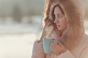 A winter's picture with girl drinking hot tea from a blue cap on a cold snowy landscape in a down jacket with a soft hood. Looking at the view. A hot steam escapes from the mug.