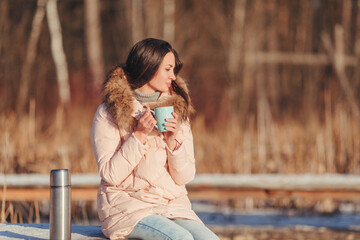 Photo of young brunette girl in a knitted sweater, pink down jacket and blue jeans. Holding a blue cup in her hands to keep warm. Standing thermos next to it. Drinking a hot tea. Looking at the side.